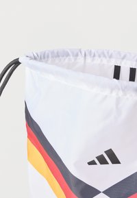 Športna torba na vrvico iz belega najlona z večbarvnimi diagonali (rdeča, rumena, črna) in črnim Adidasovim logotipom v spodnjem desnem kotu.