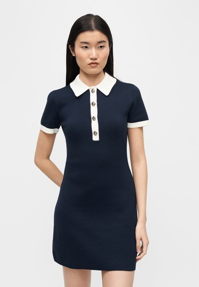 Young woman wearing a short navy dress with white collar, cuffs, and front button placket, standing against a plain light background.