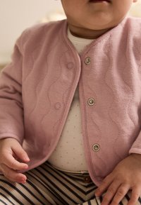 Lyserød quiltet cardigan med runde metalknapper, båret over et hvidt stykke tøj med små prikker. Kombineret med stribede bukser.