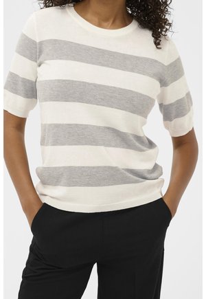 Frau trägt kurzärmeliges Strickoberteil mit breiten, horizontalen grau-weißen Streifen, kombiniert mit schwarzen Hosen, Hände in den Taschen.