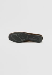 Suola di scarpa in suede marrone con un fondo in gomma nera testurizzata, caratterizzata da un motivo a onda e dettagli del logo in rilievo. Forma piatta e arrotondata.