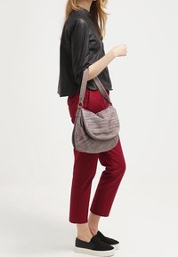 Šedá texturovaná kabelka s zakřiveným tvarem, vybavená dlouhým ramenním popruhem a zipovým uzávěrem, doplněná o červené kalhoty a černé nazouvací boty.