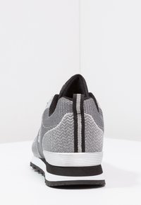 Grå träningsskor med ett texturerat meshövre, med svarta detaljer och två vertikala vita ränder på baksidan. Gummisula för bättre grepp.