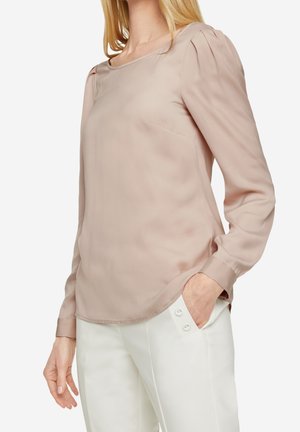 Femme portant un chemisier à manches longues rose pâle avec des épaules bouffantes et un pantalon blanc avec des détails de boutons, main dans la poche.