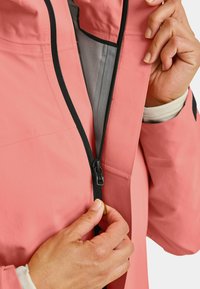 Pink waterproof jacket with a high collar, featuring a black zipper and gray lining. Hands are actively zipping the jacket.