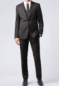 Man som bär en skräddarsydd svart tre-delad kostym med vit skjorta, svart slips och svarta finskor, stående med en hand i fickan.