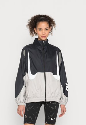 Schwarze und graue Windjacke mit hohem Kragen, elastischen Bündchen und einem weißen Logo; verfügt über Abschnitte aus glattem und strukturiertem Material.