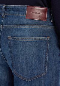 Vaqueros de mezclilla azul oscuro con un parche de cuero con el logo de Trussardi en la cintura, con costuras naranjas y un diseño clásico de cinco bolsillos.