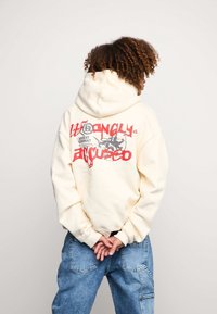 Felpa crema con un ampio design grafico rosso e grigio sul retro, polsini a coste e un cappuccio spazioso. Indossata con jeans azzurri chiari.