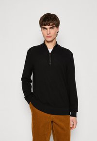 Black zip-neck pullover made from a soft material, featuring a smooth texture and long sleeves. Paired with corduroy, ribbed pants.