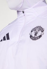 Hvit jakke med glidelås og høy krage, med en svart Manchester United-logo og tre svarte striper på venstre skulder. Glatt tekstur.