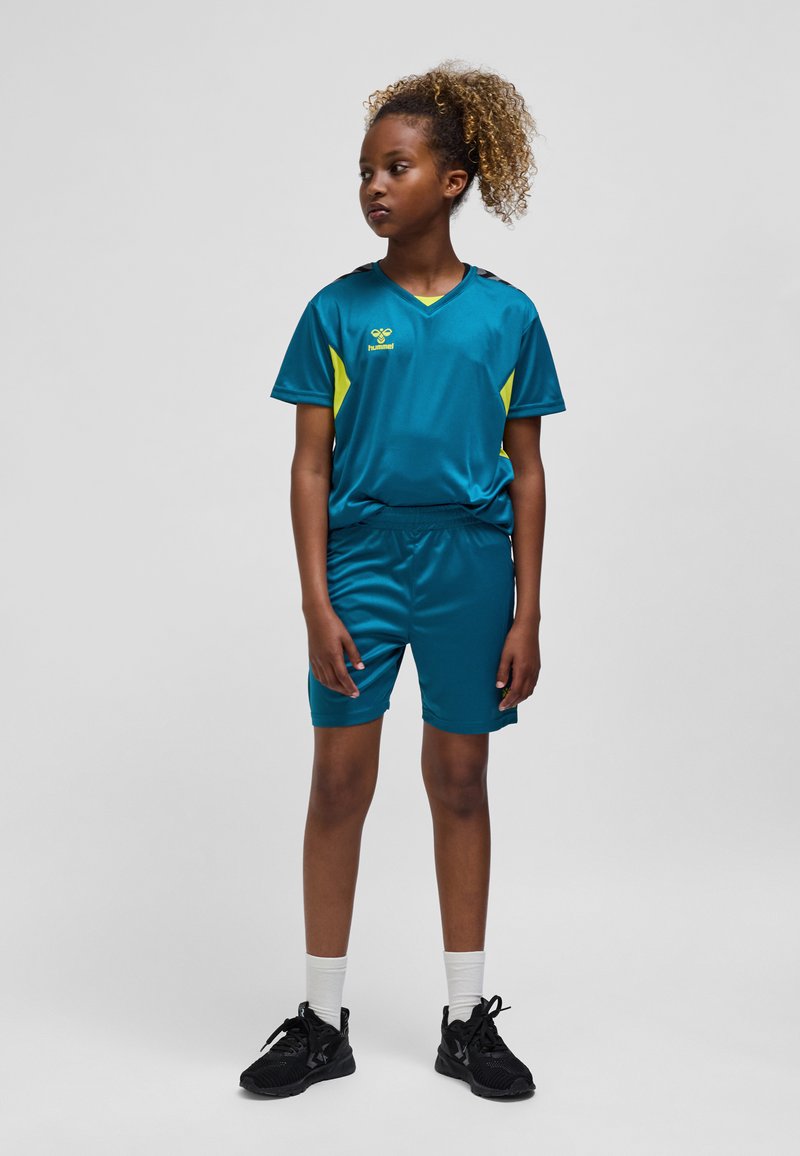 Sportstvädel för barn i blågrönt med gula detaljer, bestående av en kortärmad tröja och matchande shorts, gjorda av ett mjukt, lätt material.