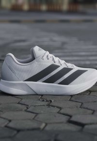 Chaussure de course Adidas Duramo RC 2 blanche avec des bandes noires sur une surface pavée en pierre grise.