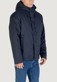 Giacca insulata blu navy con cappuccio, dotata di tasche laterali con zip e un esterno liscio e resistente all'acqua, progettata per calore e comfort.