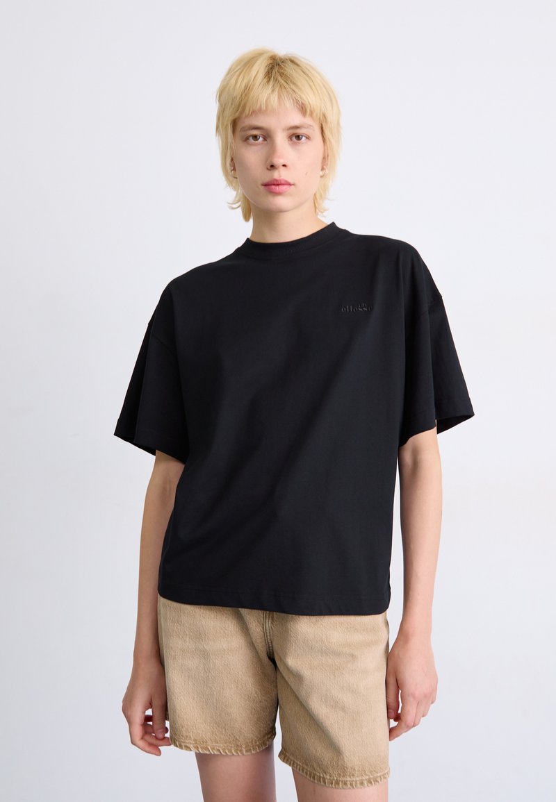 Maglietta nera oversize in cotone, con scollo rotondo e maniche corte. Dettaglio del logo goffrato sul petto.