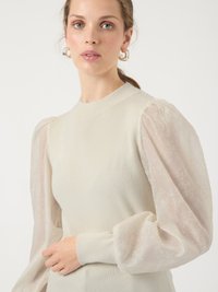 Woman wearing beige ribbed sweater with sheer puffed floral embroidered sleeves and hoop earrings, against a plain light background.