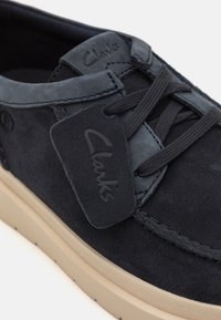 Dark blue suede shoe featuring a rubber sole, round laces, and a branded tag with 'Clarks.' The design is casual and modern.