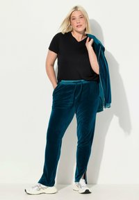 Fluwelen trainingspak in teal, met een zwart V-hals t-shirt. De broek heeft zijsplitsen en een tailleband met trekkoord. Witte sportschoenen maken de look compleet.