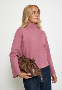 Ružový sveter s mock turtleneck, dlhými rukávmi, skombinovaný s hnedou koženou taškou na šnúrku s zhlčkovaným detailom a textúrovaným povrchom.