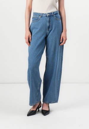Model in a beige top and blue wide-legged jeans, standing on a white background, wearing black pointed-toe heels.