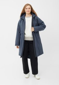 Mørkeblå lang pufferjakke med hætte, lynlås og elastisk snøring i taljen. Båret over en lys grå sweatshirt og sorte bukser.
