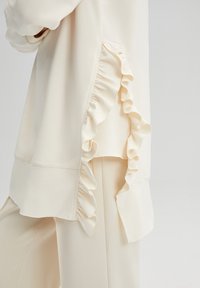 Cream-colored top with a ruffled detail along the side seam, featuring a smooth texture and a relaxed fit.