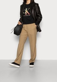 Blazer in pelle nera sopra una felpa nera con logo, pantaloni color cammello e sneaker nere con suole bianche, che tiene una borsa nera.