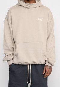 Sweat à capuche beige surdimensionné avec une poche kangourou, des cordons de serrage et un logo discret sur la poitrine ; confectionné dans un tissu doux et texturé.