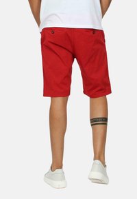 Shorts rossi in cotone con due tasche posteriori con bottoni marroni, abbinati a una maglietta bianca e sneaker grigie chiaro. Tatuaggi visibili sul polpaccio.