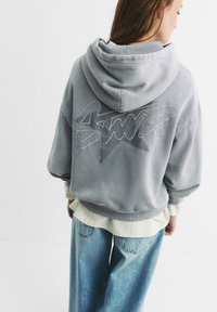 Un hoodie gris surdimensionné avec un logo et un design d'étoile bien en vue au dos. Il possède une texture douce et une coupe décontractée, associé à un jean bleu ample.