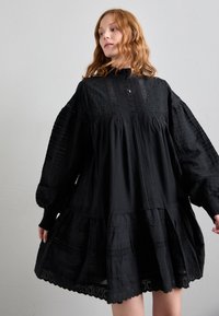 Črna plise obleka z visokim ovratnikom, dolgimi napihnjenimi rokavi, teksturiranim materialom, gumbno zapenjanjem in čipkastim robom.