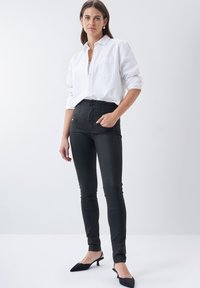 Zwarte high-waisted jeans met een gladde textuur, gecombineerd met een witte blouse met knopen. Het model draagt zwarte pumps met een spitse neus.