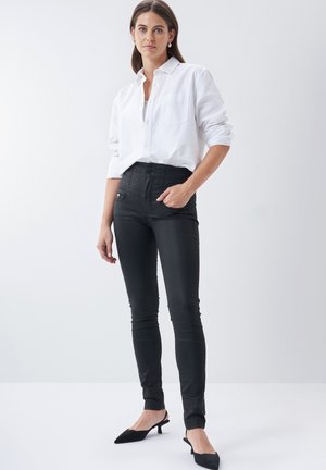 Jeans negros de cintura alta con una textura suave, combinados con una camisa blanca de botones. La modelo lleva puestos unos tacones negros de punta.