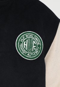 Czarny i kremowy polarowy sweatshirt z okrągłą, zieloną haftowaną naszywką z napisem "HUF WORLDWIDE TRANSIT AUTHORITY" oraz szczegółami logo.