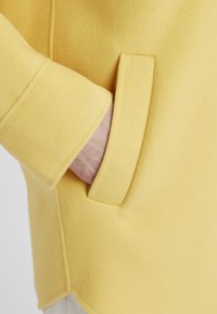 Gelber Wollmantel mit Seitentasche und glatter Textur, leicht geschwungem Saum und strukturiertem Kragen. Minimalistisches Design.