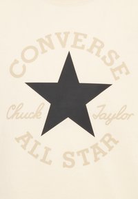 Kremowa koszulka z dużą czarną gwiazdą, z napisem "CONVERSE" i "ALL STAR" w beżowym kolorze oraz "Chuck Taylor" w kroju pisma kursywą.