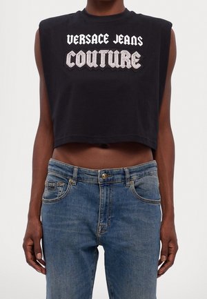 Personne portant un haut court noir sans manches avec le texte "Versace Jeans Couture" et un jean denim bleu avec un patch de marque sur la poche.