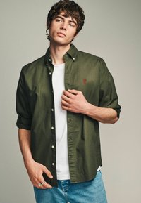 Chemise boutonnée vert olive en coton, avec un petit logo rouge sur la poitrine, superposée à un T-shirt blanc, accompagnée d'un jean bleu.