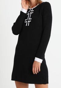 Žena v černých dlouhých šatech s bílým lemem na manžetách a výstřihu, které mají dekorativní bílé pruhy s mašlemi na hrudi.