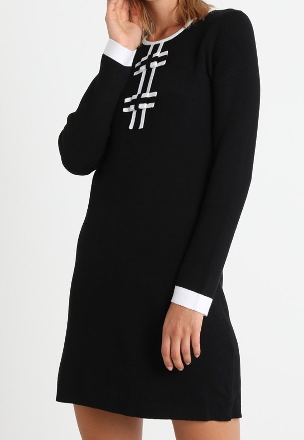 Žena v černých dlouhých šatech s bílým lemem na manžetách a výstřihu, které mají dekorativní bílé pruhy s mašlemi na hrudi.