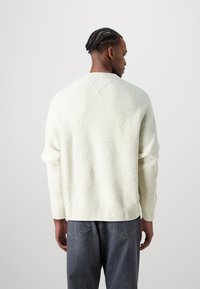Suéter de punto color crema con corte relajado, puños y dobladillo acanalados, y un yugo trasero en forma de V, combinado con jeans oscuros.