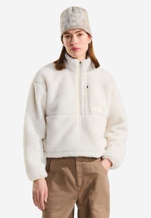 Ung kvinde iført en hvid fleece jakke med lynlås, beige mønstret hue og brune bukser står med den ene hånd i lommen.