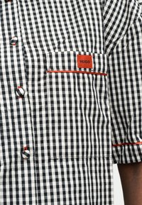 Πουκάμισο με ασπρόμαυρο καρό και κόκκινο τελείωμα, κουμπιά και τσέπη στο στήθος με ετικέτα "HUGO" στο πάνω μέρος του μανικιού.