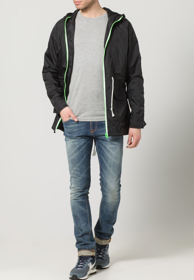 Svart lett jakke med neon grønne detaljer, glidelås og to frontlommer, paret med grå T-skjorte og blå jeans.
