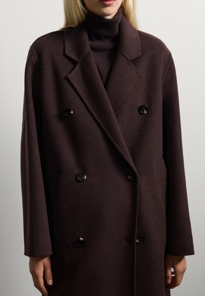 Femme portant un manteau en laine marron foncé à double boutonnage et un pull col roulé assorti, se tenant devant un fond de mur clair et uni.