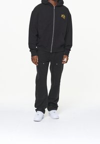 Felpa nera con zip e tasche frontali, con logo dorato, abbinata a jeans ampi neri con tasche cargo, completata da sneakers bianche.