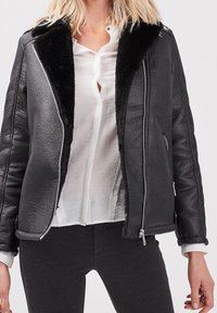 Černá kožená bunda s límcem z umělé kožešiny, zapínáním na zip, prošívanou strukturou a bočními kapsami. Nošená přes bílou košili na zapínání.