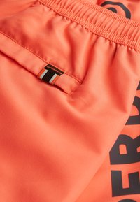 Pantaloni sportivi arancioni realizzati in un tessuto liscio e elasticizzato, con cintura elasticizzata e un'etichetta con logo caratterizzata da strisce nere e arancioni.