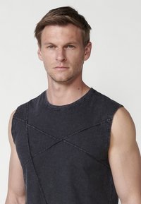 T-shirt en coton noir sans manches, ornée d'un motif de couture croisée sur la poitrine et de bords bruts, avec un col rond et une coupe décontractée.