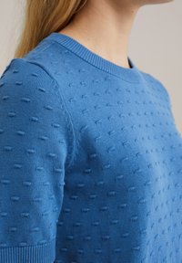 Pītā zila džemperis ar teksturētu rakstu un maziem paceltiem punktiem, apļveida kakls un ribotas aproces. Gluda auduma apdare.